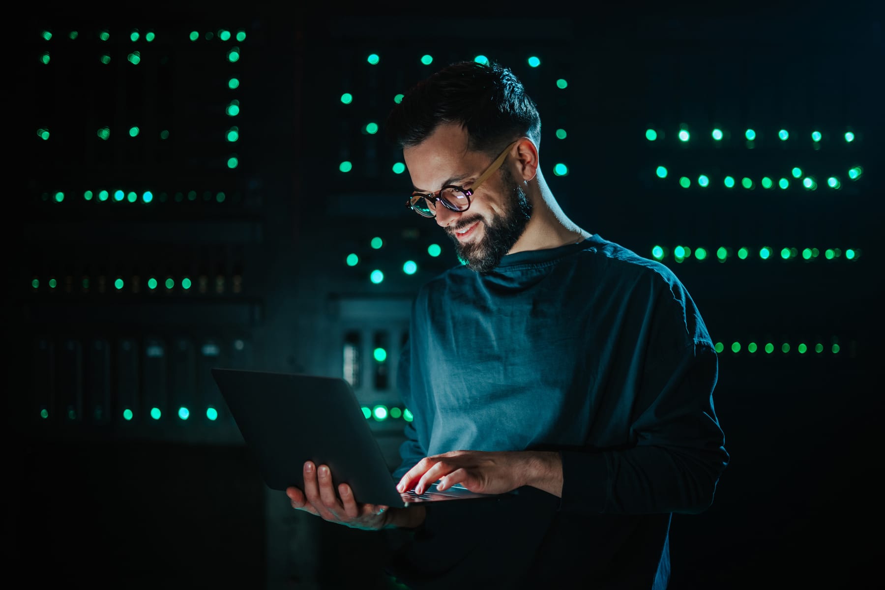 Engineer working on a laptop in a dark server room with rows of green LED lights.