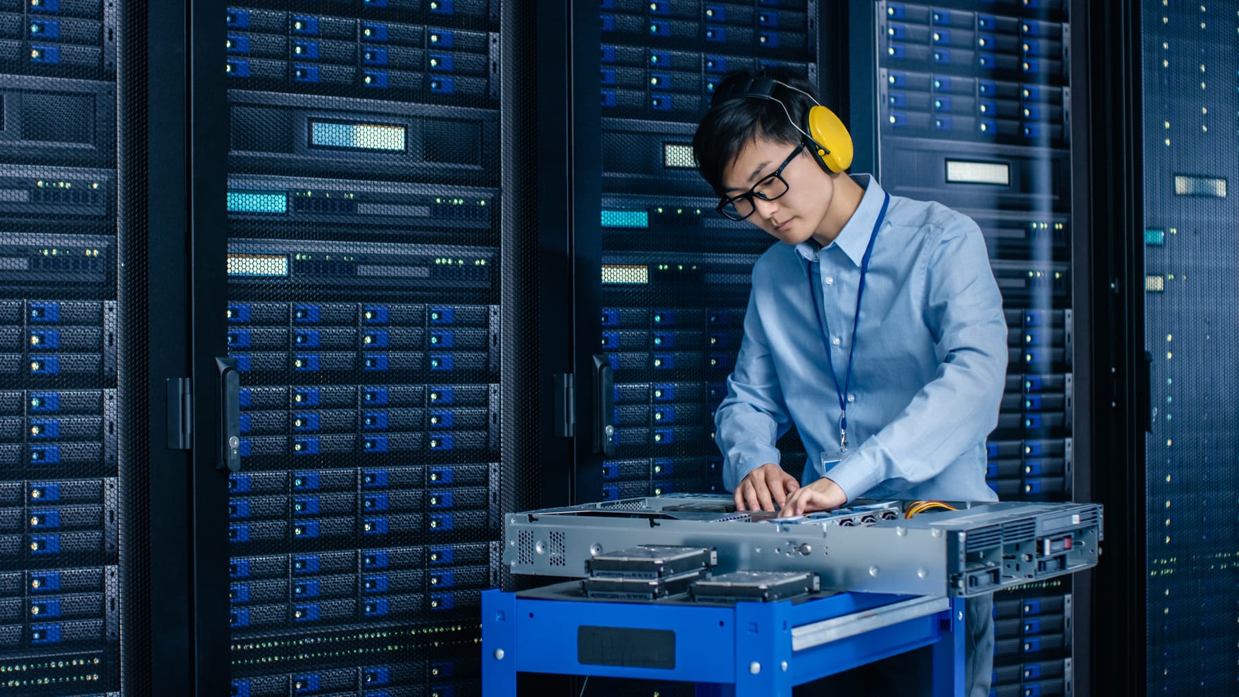 Technician with ear defenders working on an open server on a cart beside dark racks.