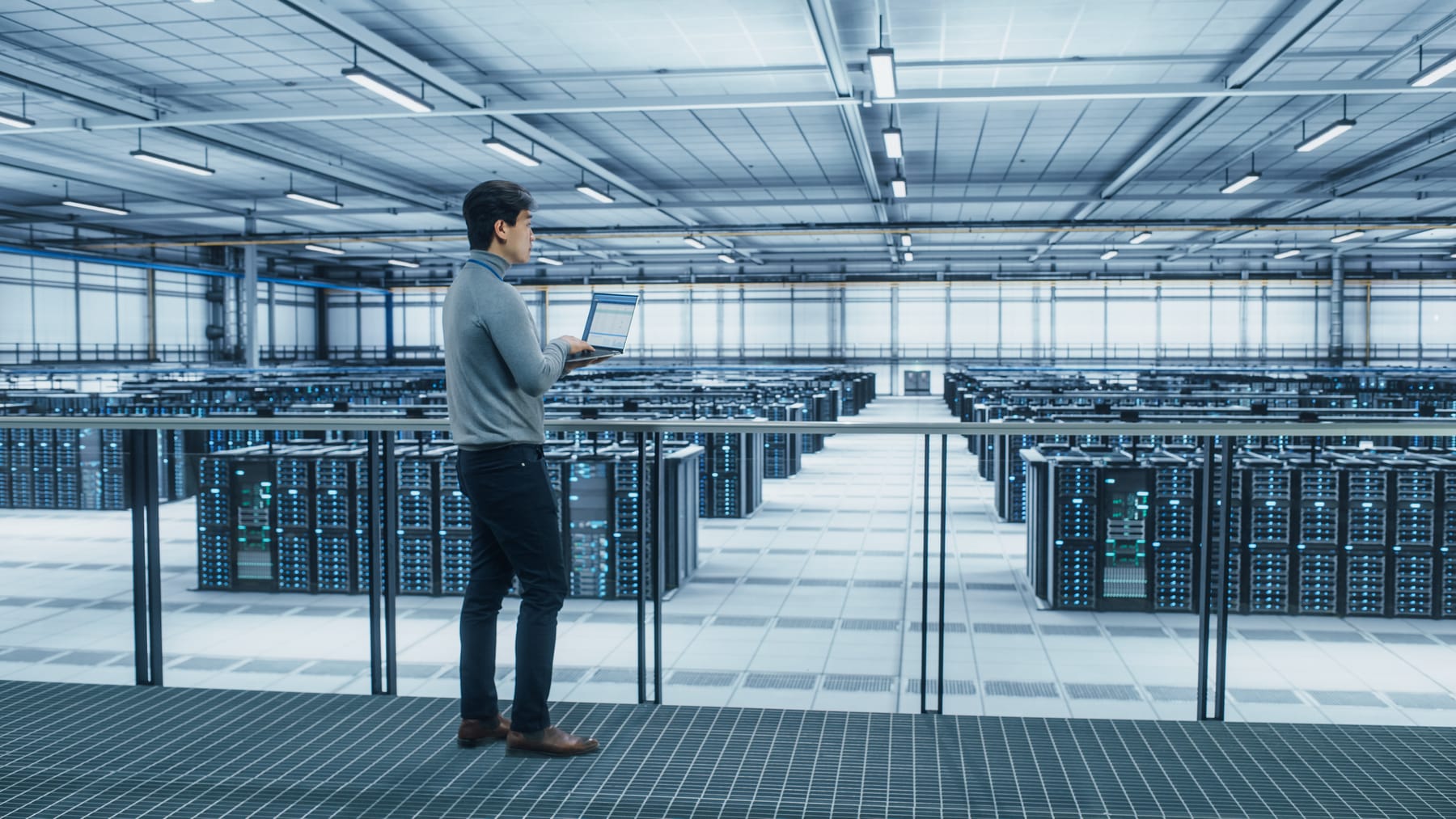 Engineer with a laptop on an elevated walkway in a bright modern hyperscale data hall.