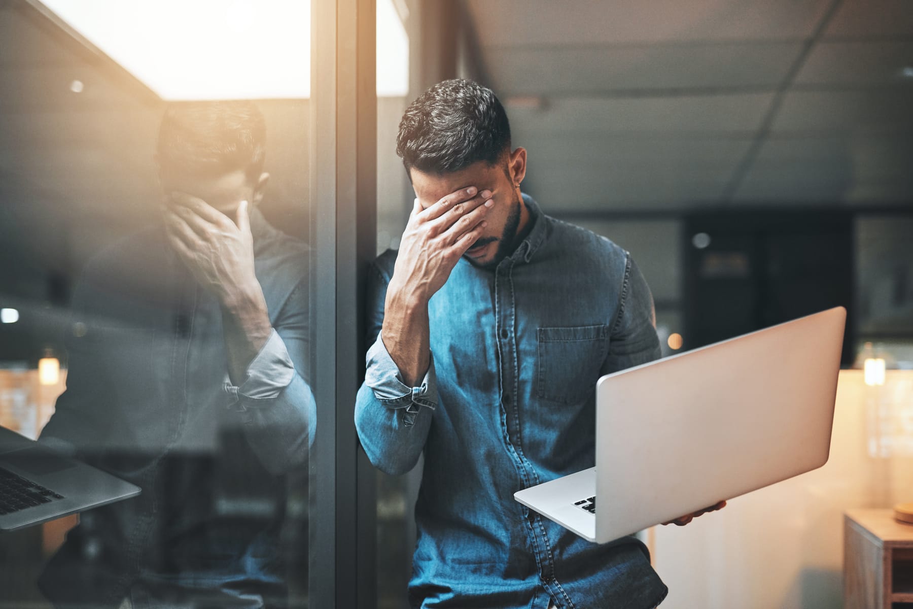Frustrated man holding a laptop with his hand over his face, reflected in office glass.