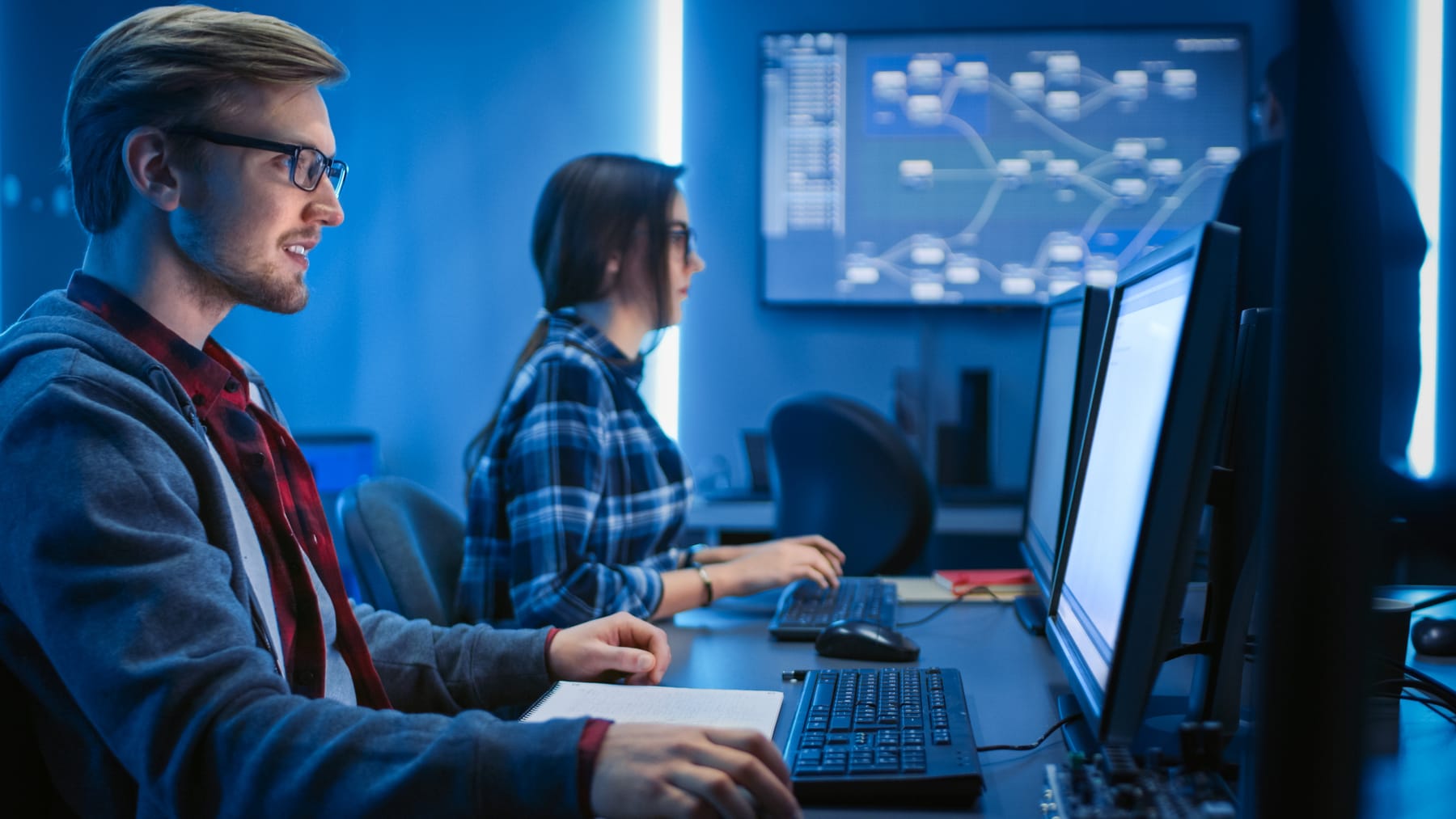 Developers working at desks with a large network topology diagram on a wall screen.