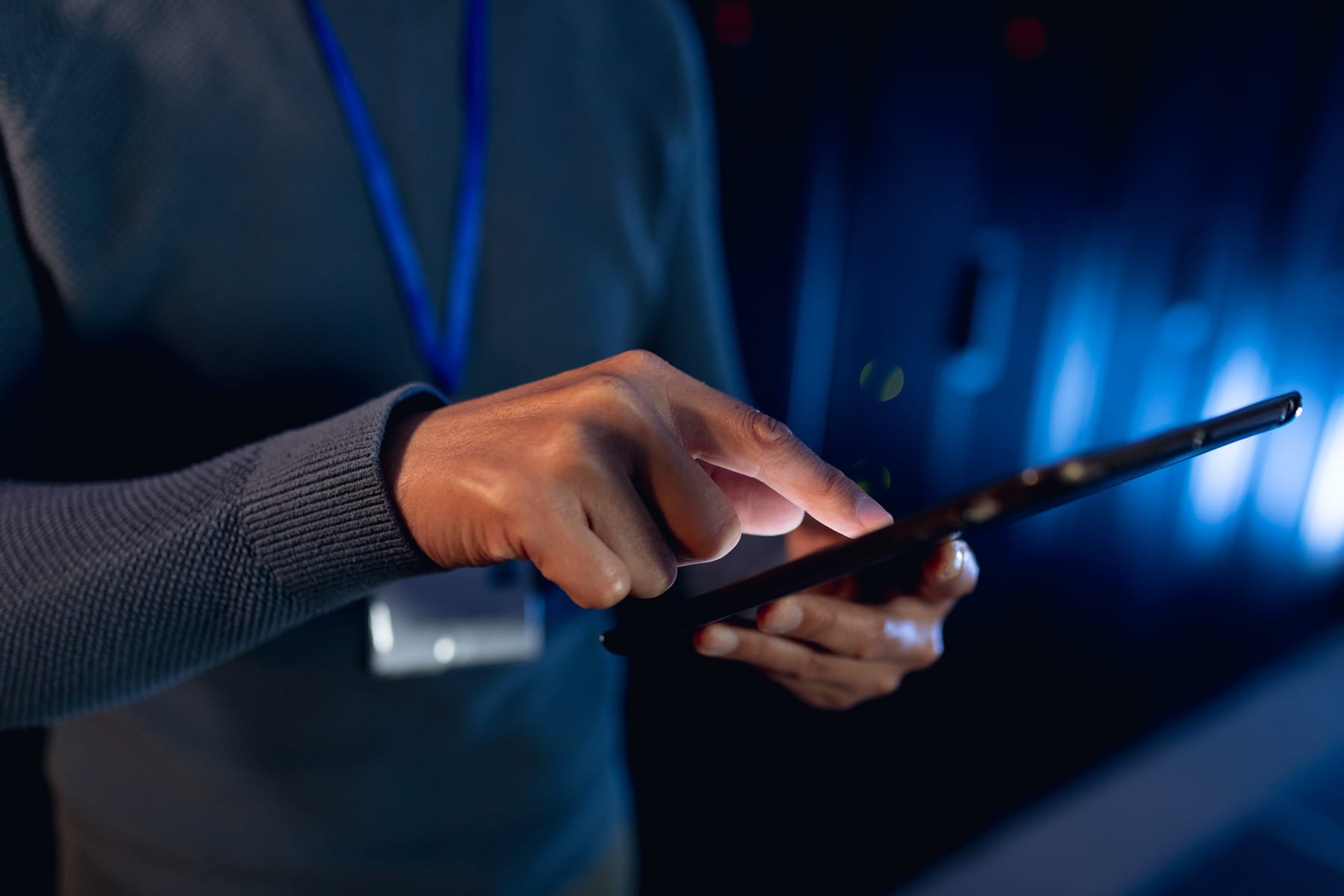 Close-up of hands using a tablet in a dark server room with blue rack lights behind.