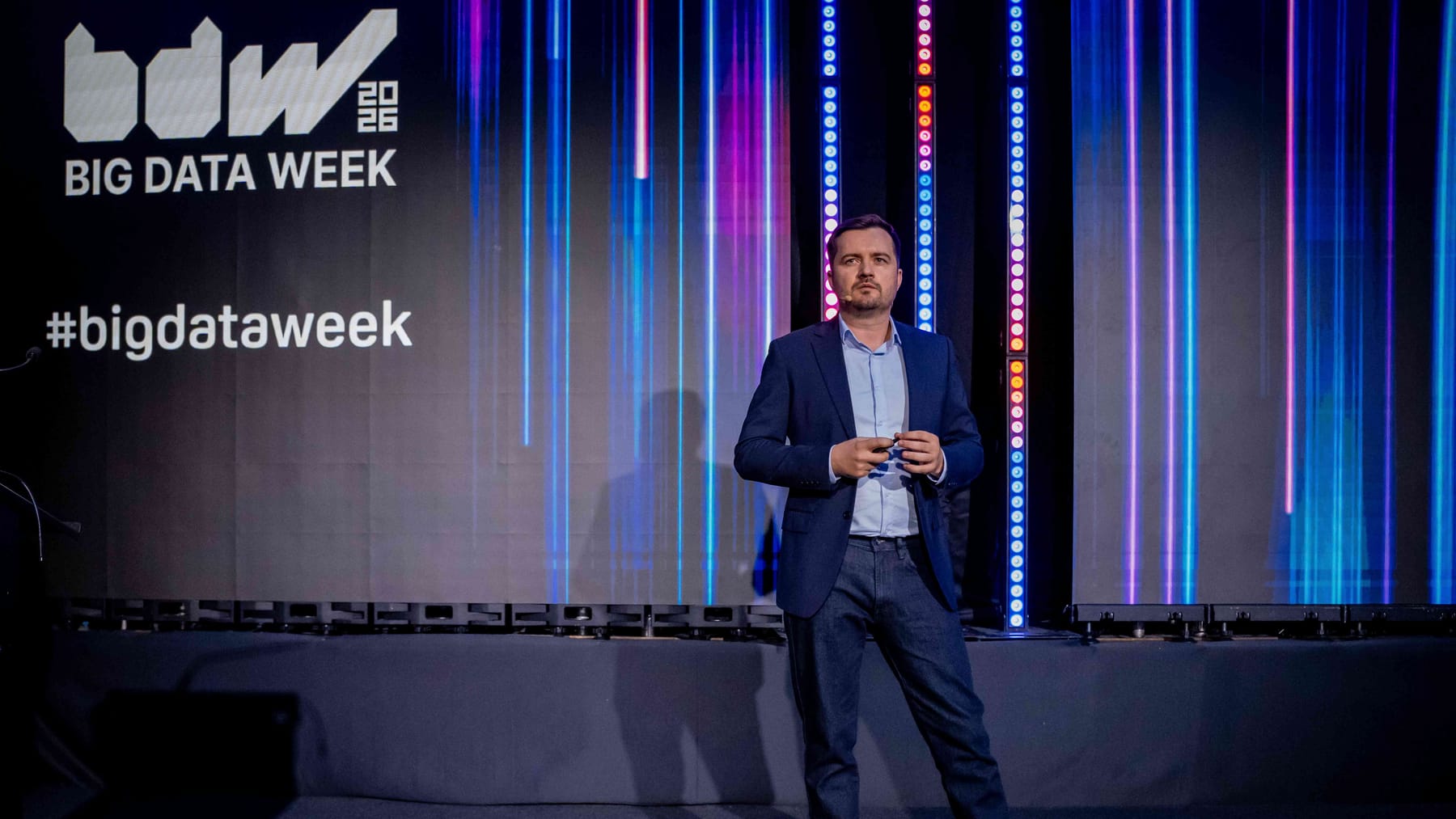 Andrei Tomus speaking on the Big Data Week Bucharest 2026 stage, in front of a dark screen showing the conference logo, the #bigdataweek hashtag and vertical streaks of blue and pink light.