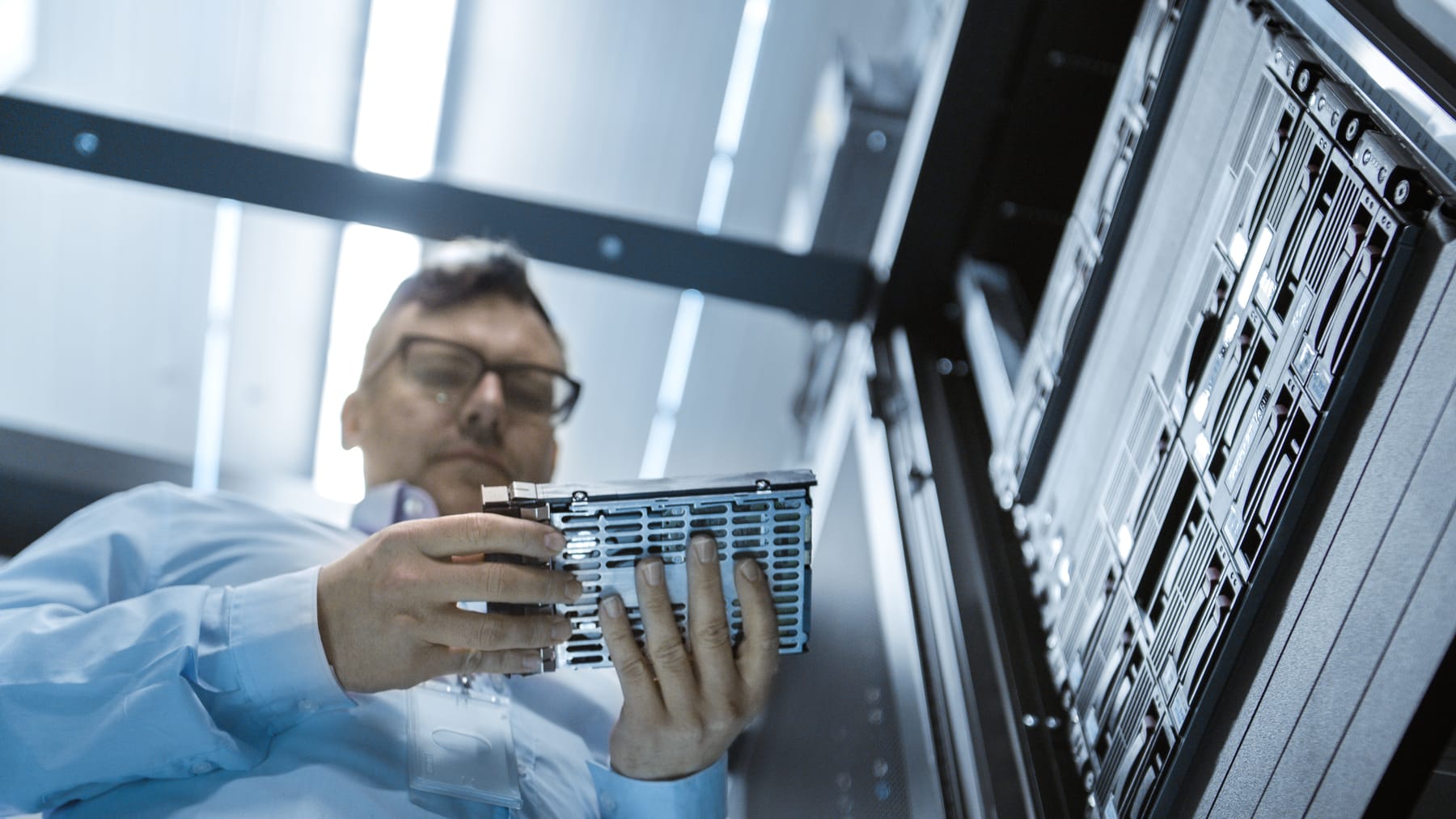 Technician examining a hard drive in its caddy beside an open storage rack.