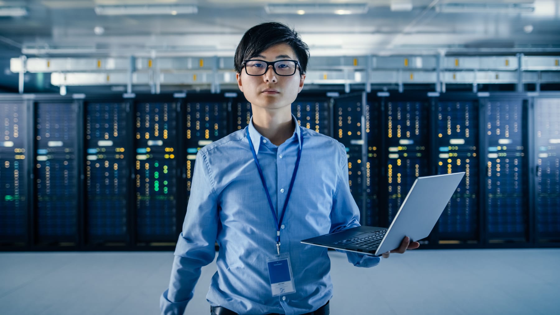 Technician in glasses holding a laptop in front of server racks with amber and green LEDs.