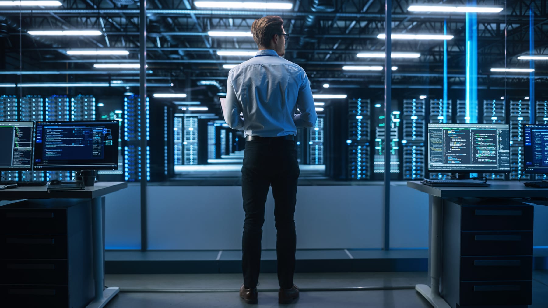 Engineer seen from behind standing between monitors, facing long rows of illuminated racks.