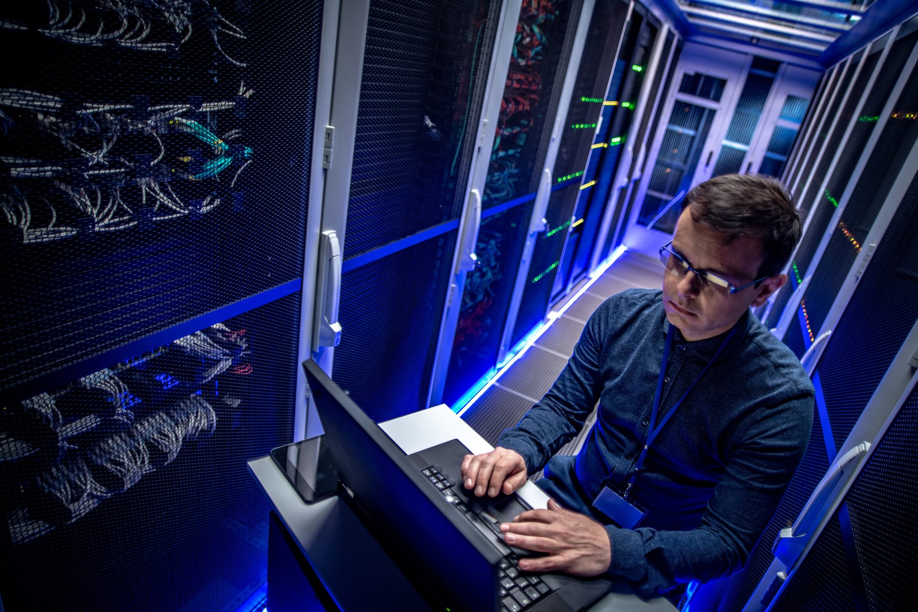 Engineer typing on a laptop atop a cart in a dramatically blue-lit server aisle.