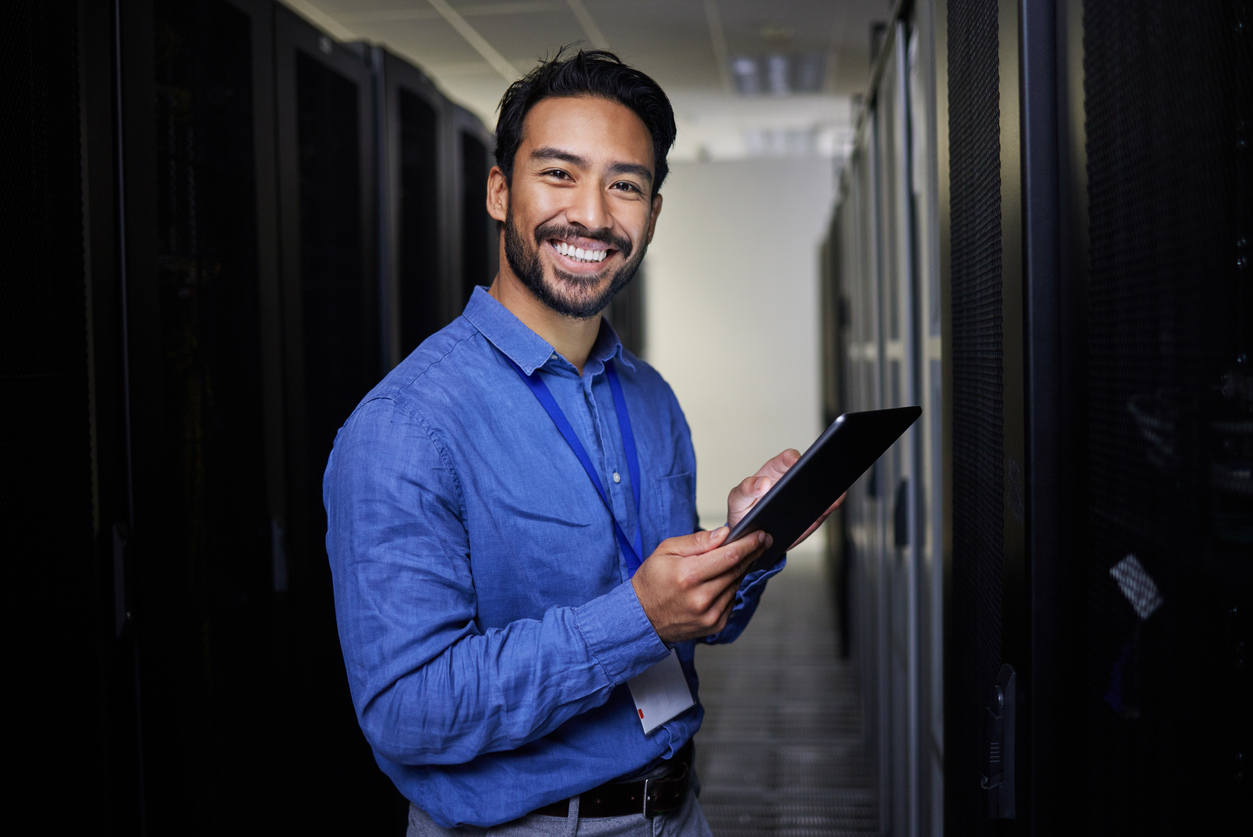 The same IT administrator standing in a bright server aisle, smiling confidently while working on a tablet